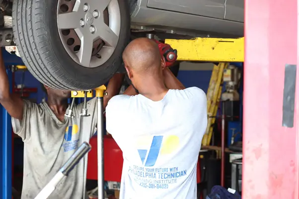 Two PTTI auto technicians raise a transmission jack under a lifted car in a repair shop.